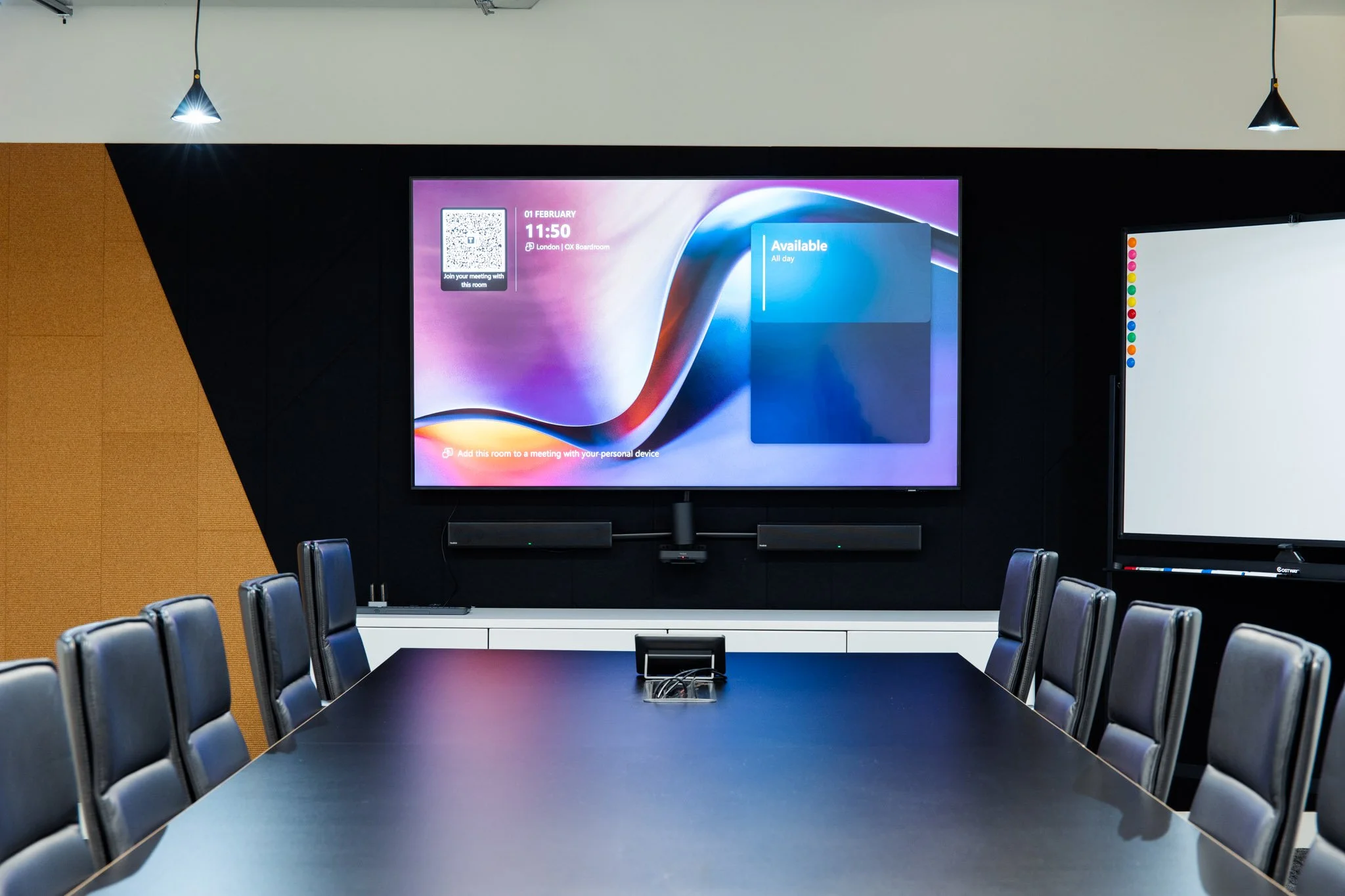 Conference room with a large black table surrounded by black chairs, a wall-mounted digital display showing meeting details, and a whiteboard on the side.