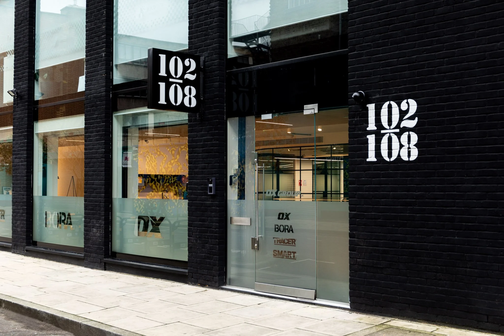 Modern storefront with large glass windows and black brick facade, displaying the number 10208 on a black sign and white logo on the glass.