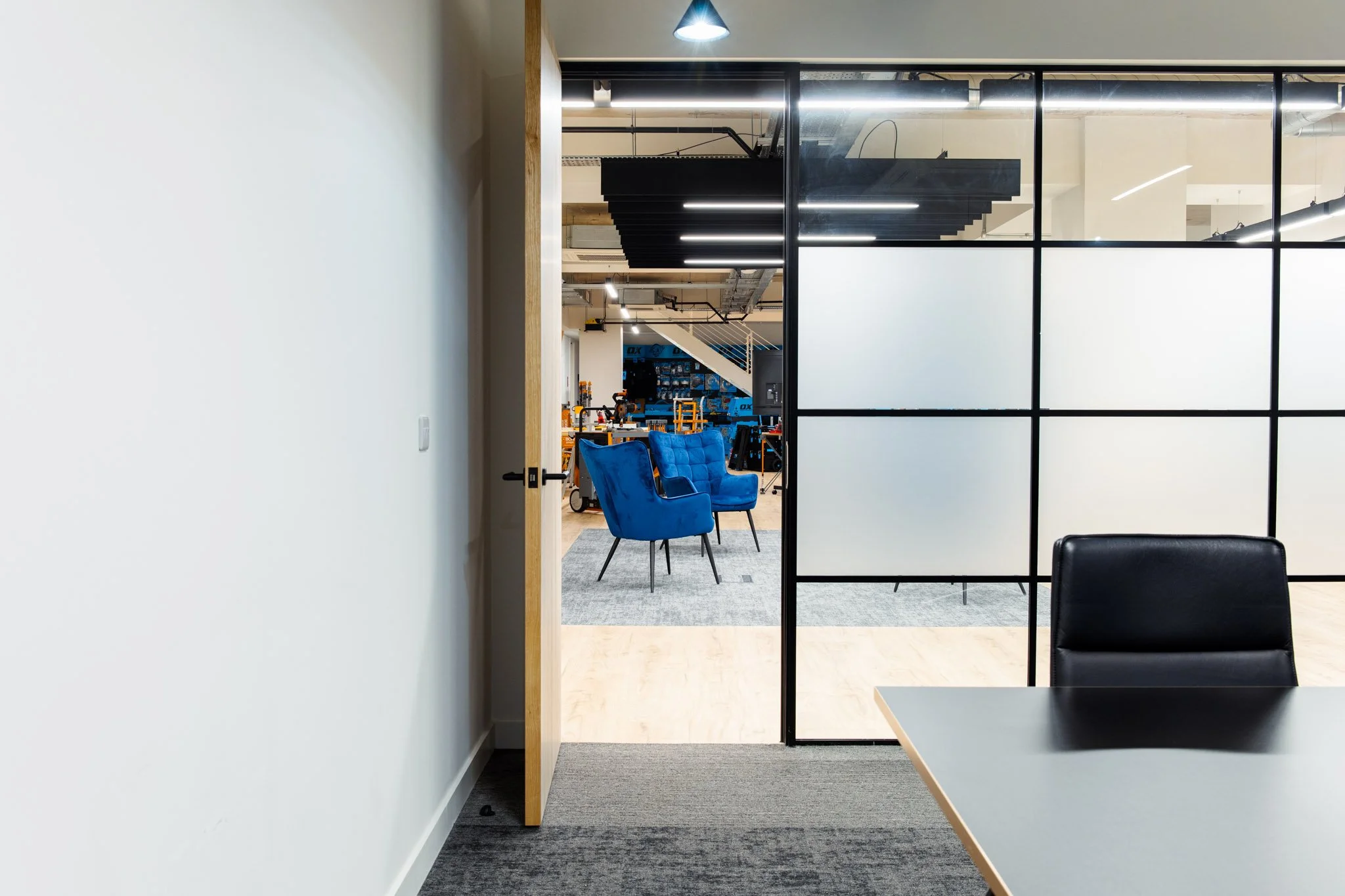 The image shows a modern office or workspace seen through a glass partition. Inside, there are blue chairs and a table in the foreground, and a work area with shelving, tools, and equipment in the background. Overhead lighting and a staircase are vis