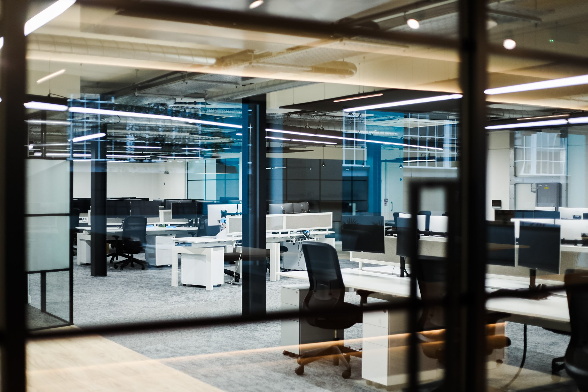 A modern office interior viewed through a glass window with blinds, featuring desks, chairs, and computer monitors.