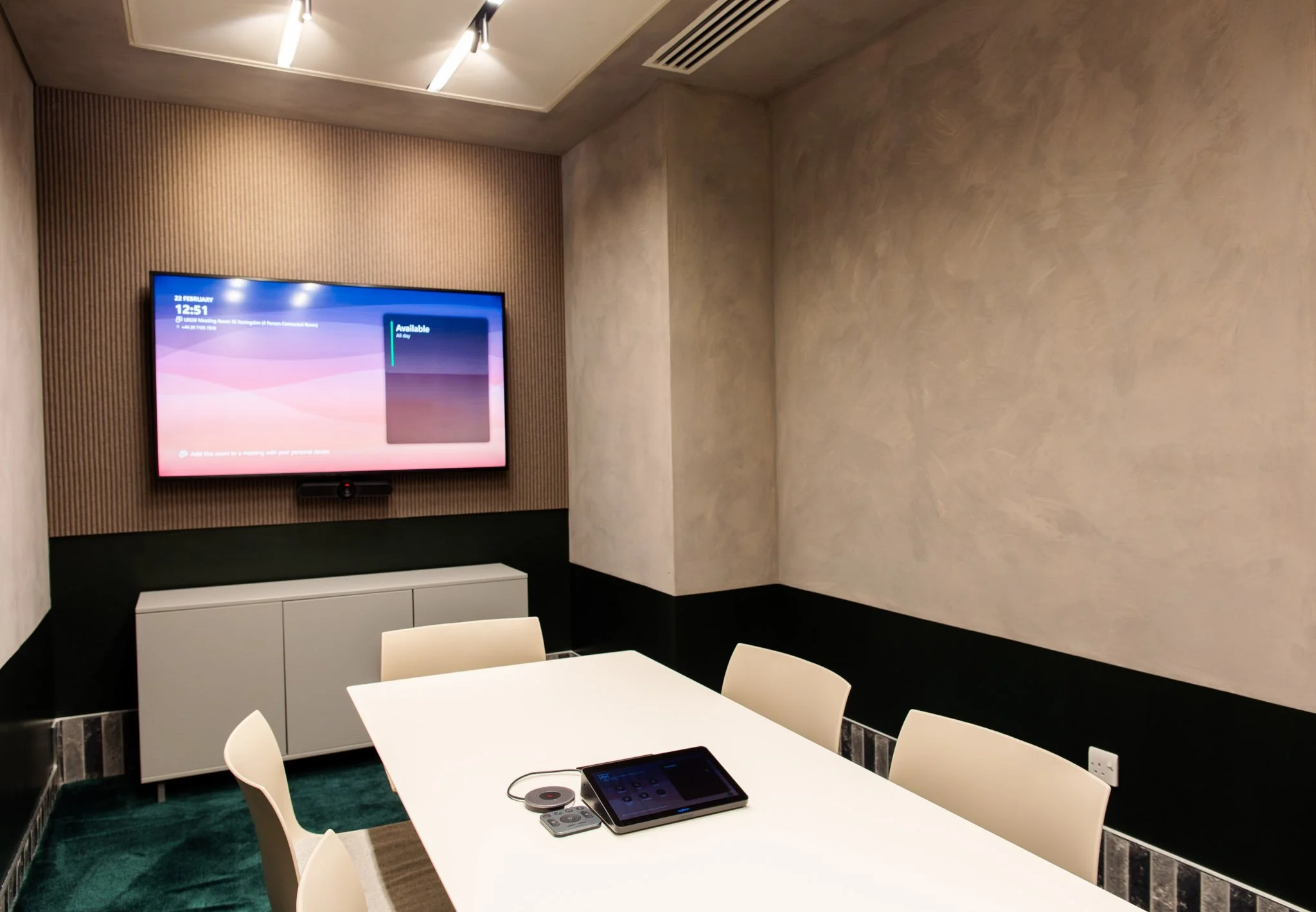 A conference room with a white table, beige chairs, a wall-mounted flat-screen monitor displaying a schedule, and a conference phone on the table. The room has beige textured walls, a white cabinet, and a ceiling with modern lighting and an air vent.