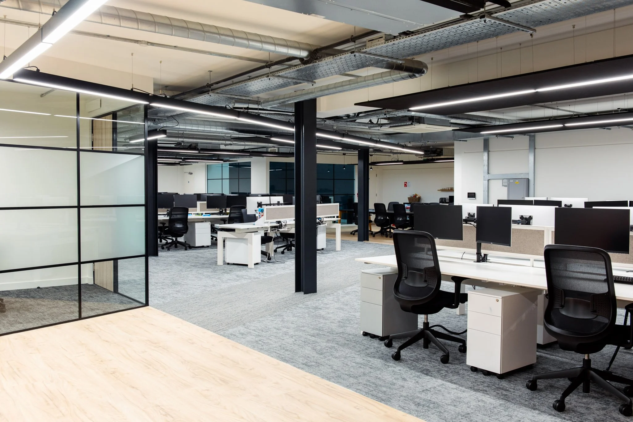 Modern open-plan office with white desks, black office chairs, multiple monitors, and exposed ductwork on the ceiling.