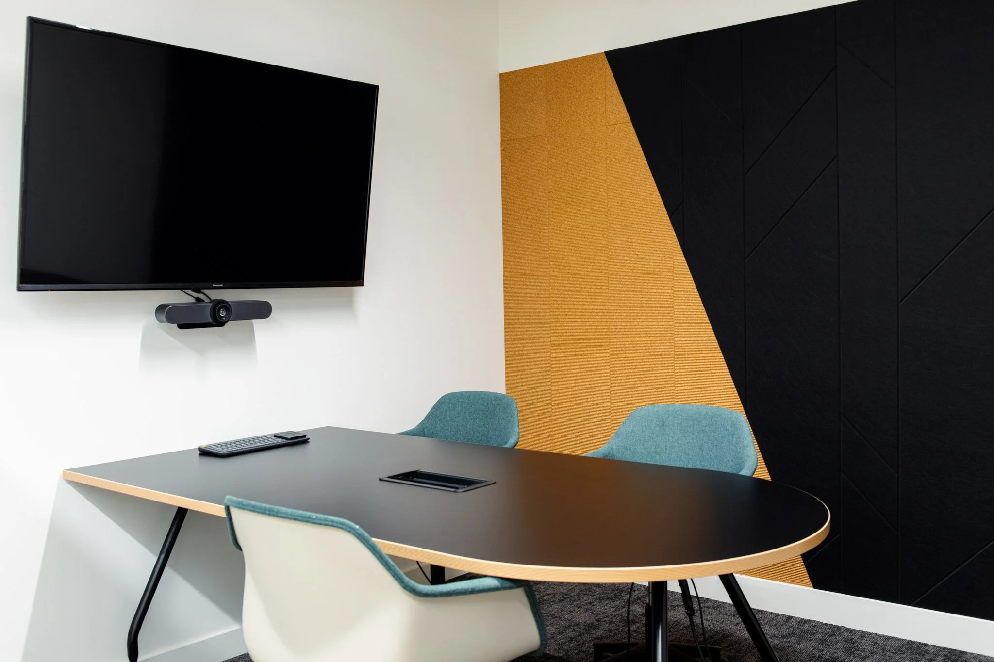 Modern meeting room with a large television, a conference table with a remote and keyboard, colorful chairs, and a textured wall with black and mustard sections.