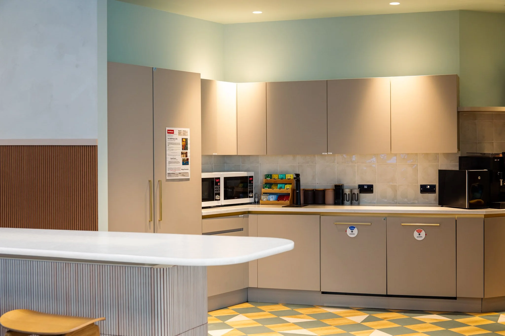 A modern kitchen with beige cabinets, a microwave oven, and a coffee station, with a yellow and gray checkered floor and light green walls.