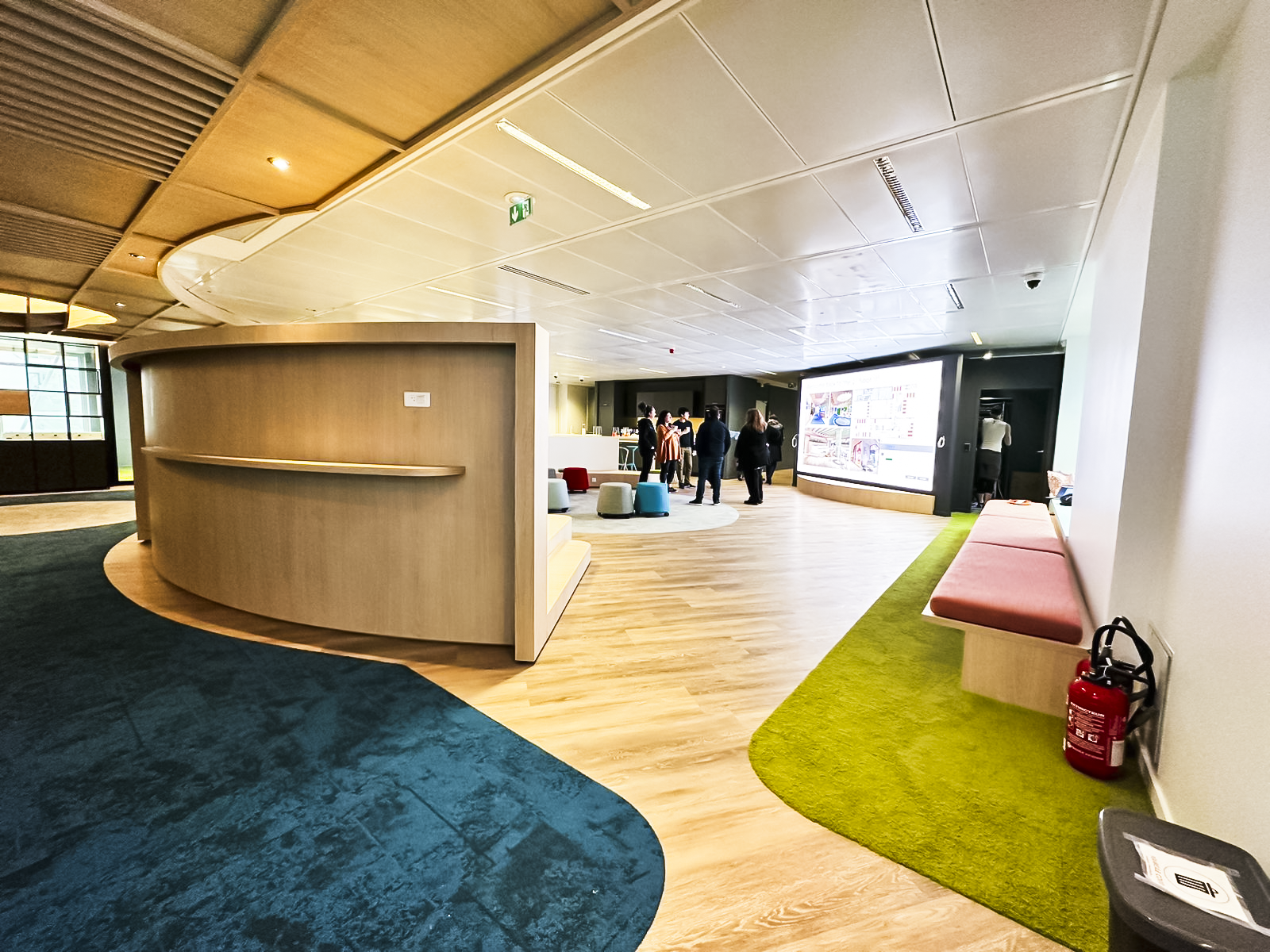 Modern indoor lobby area with curved wooden reception desk, colorful seating, large digital display screen, and people gathered in the background.