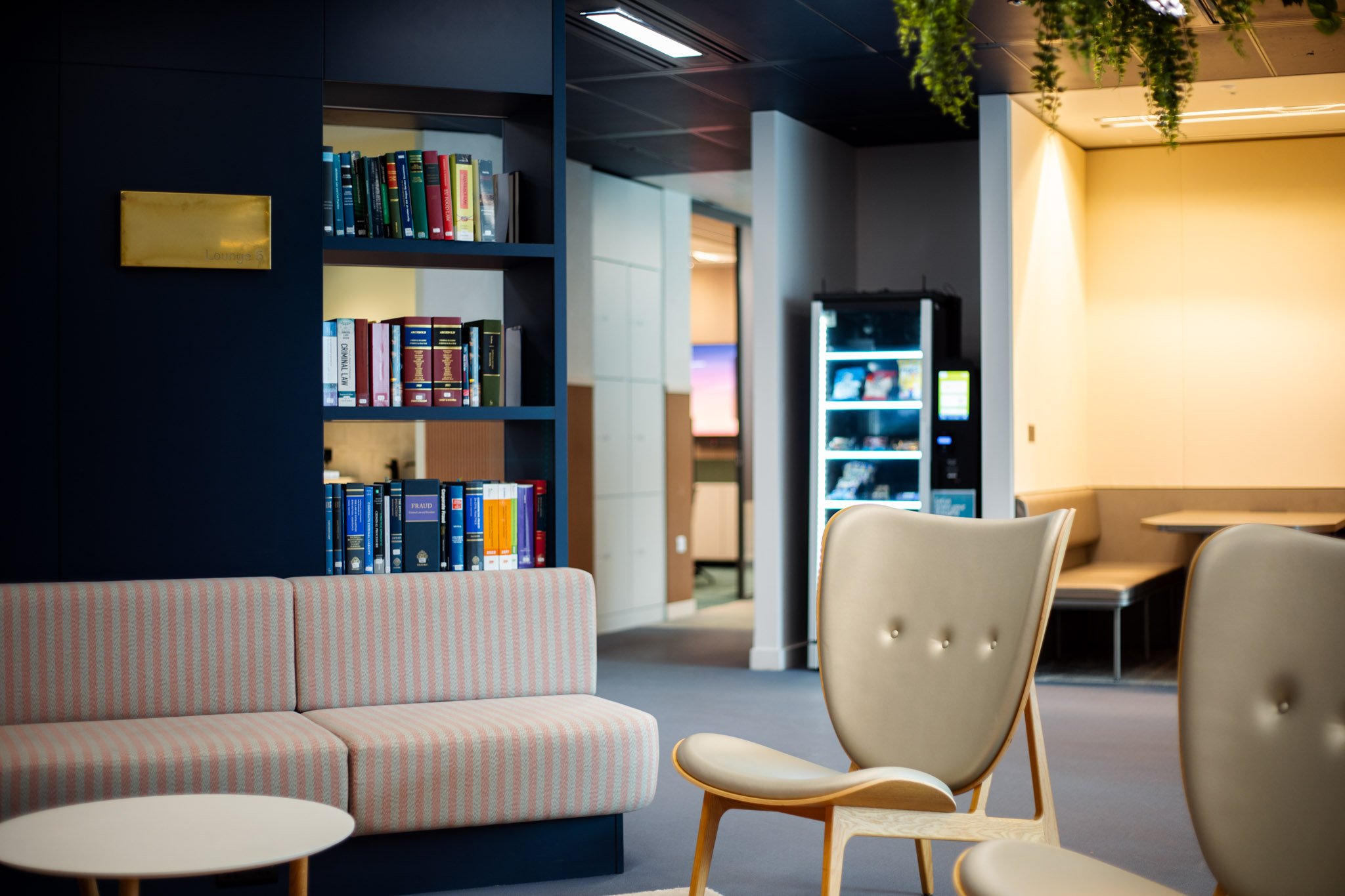 Interior of a lounge area with beige chairs, a pink striped sofa, books on dark shelves, and a vending machine against a light-colored wall. Green plants hang from the ceiling.