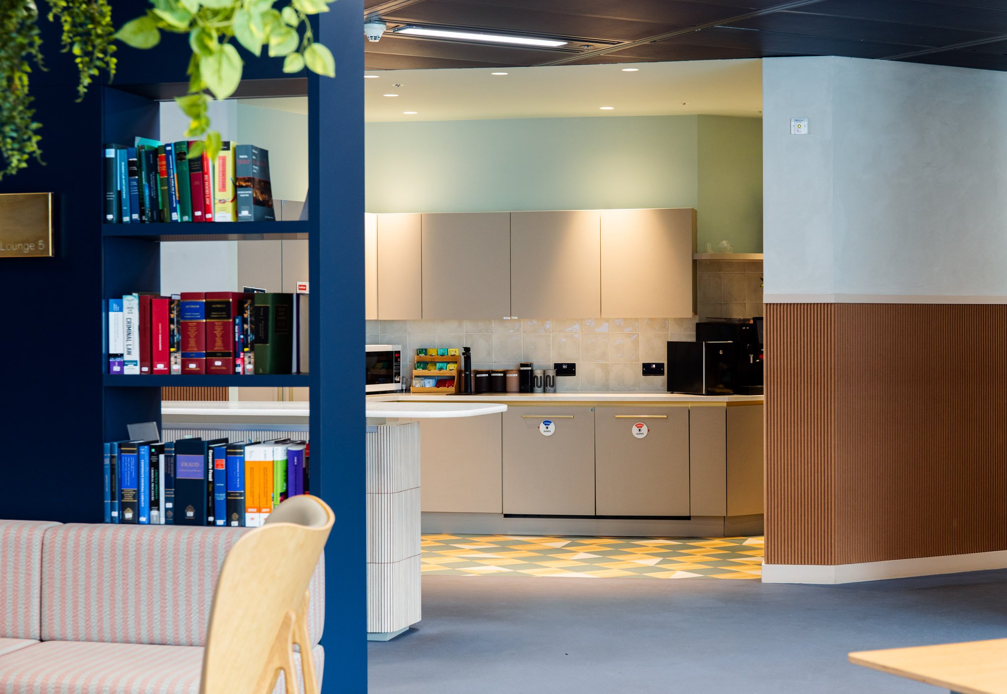 Interior of a modern cafe or lounge area with a bookshelf, a seating area with a patterned cushion, and a kitchen or service area with cabinets and appliances in the background.