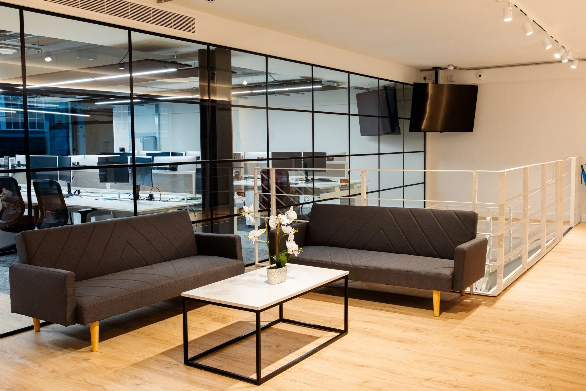 Modern office lounge with two brown sofas, a white marble coffee table with a small plant, a glass wall showing an open office space with desks and computer monitors, and a wall-mounted flat-screen TV.