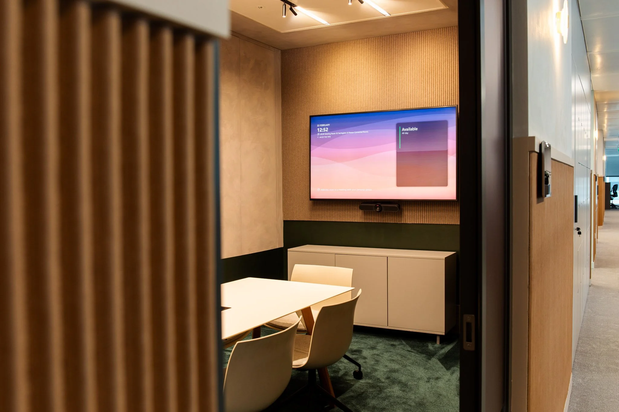 A conference room with a large wall-mounted screen displaying the date, time, location, and availability; partially visible table and chairs in the foreground, and hallway lighting in the background.