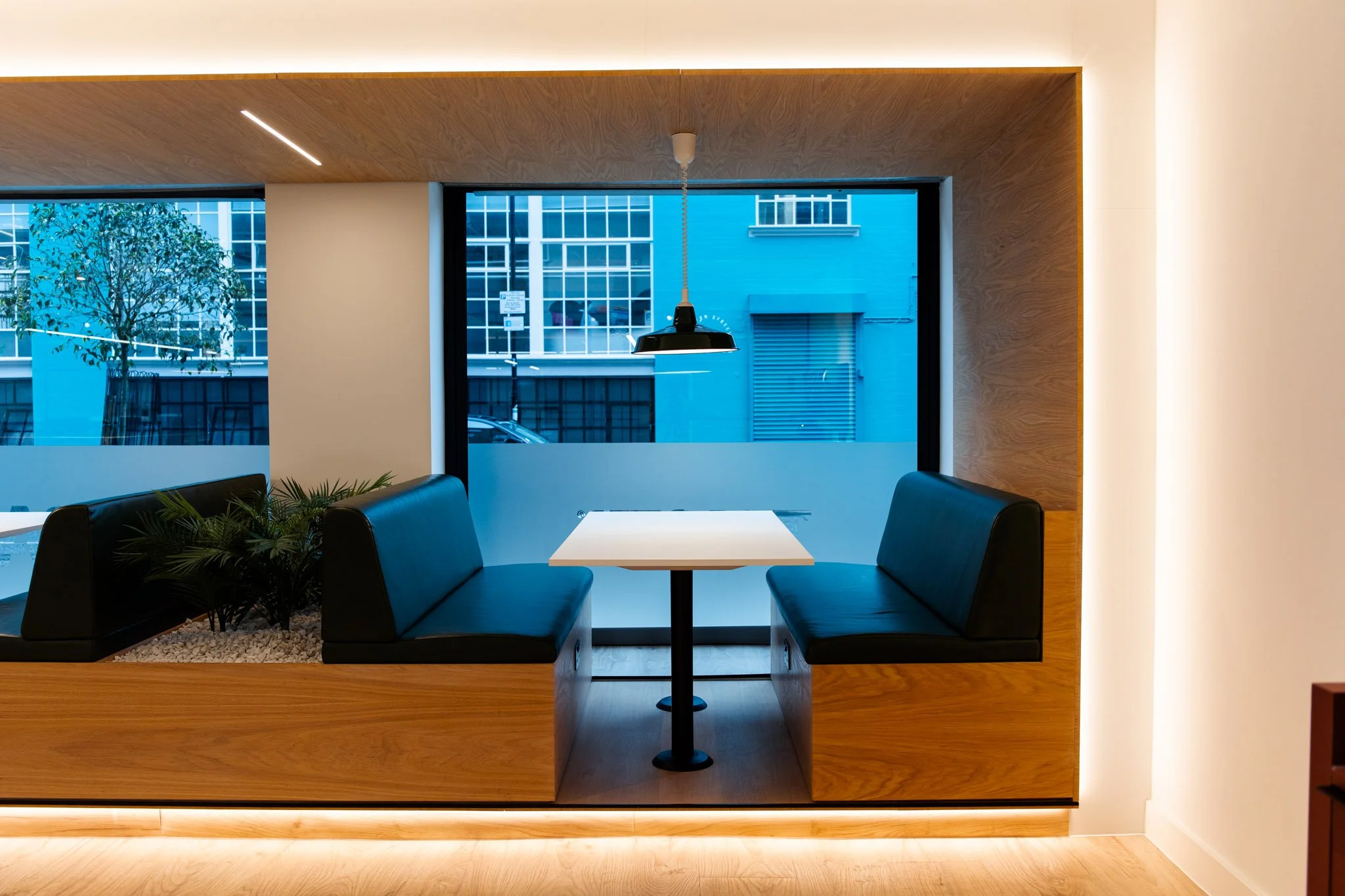 Interior of a modern dining area with a white table and black cushioned benches. Large window with a cityscape view, wooden accents, and a pendant light hanging above the table.