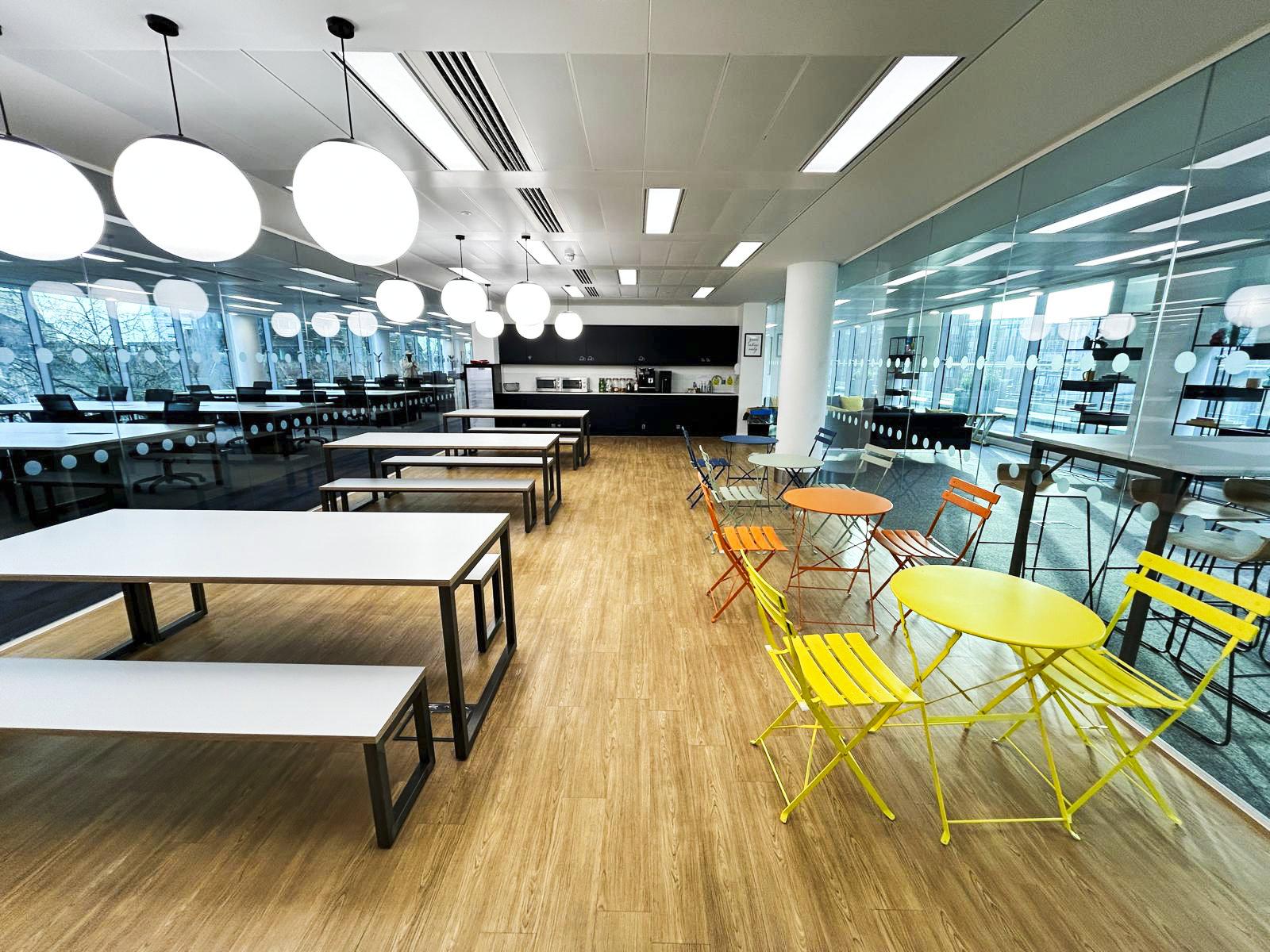 A modern, empty cafeteria with white tables, colorful chairs, and large windows.