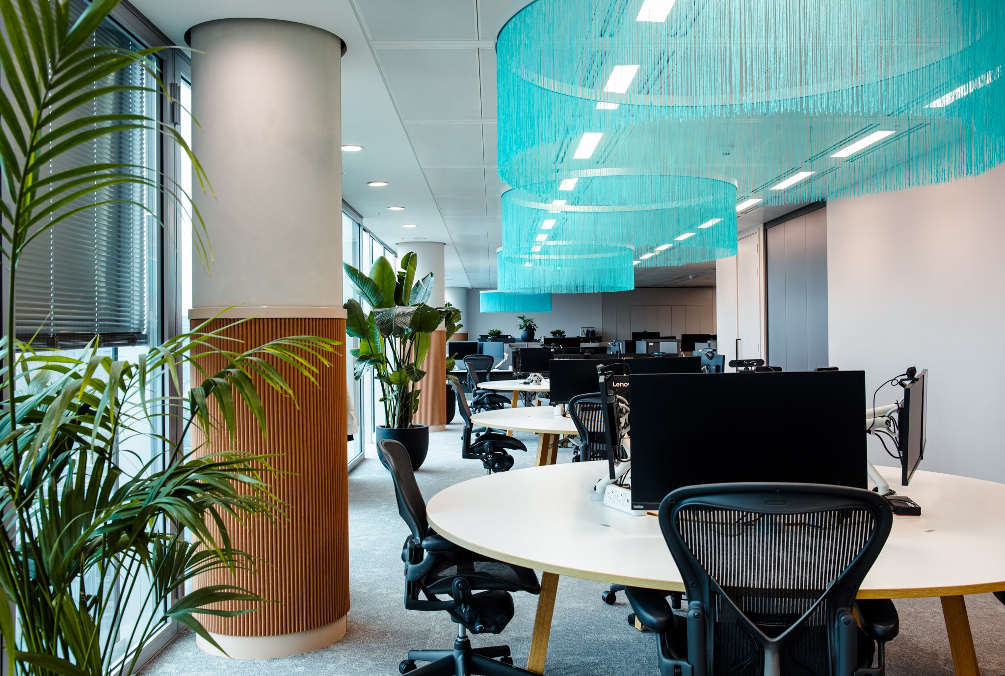 Modern office space with round white tables, black office chairs, several computer monitors, large windows, indoor plants, and a large blue hanging chandelier ceiling decor.