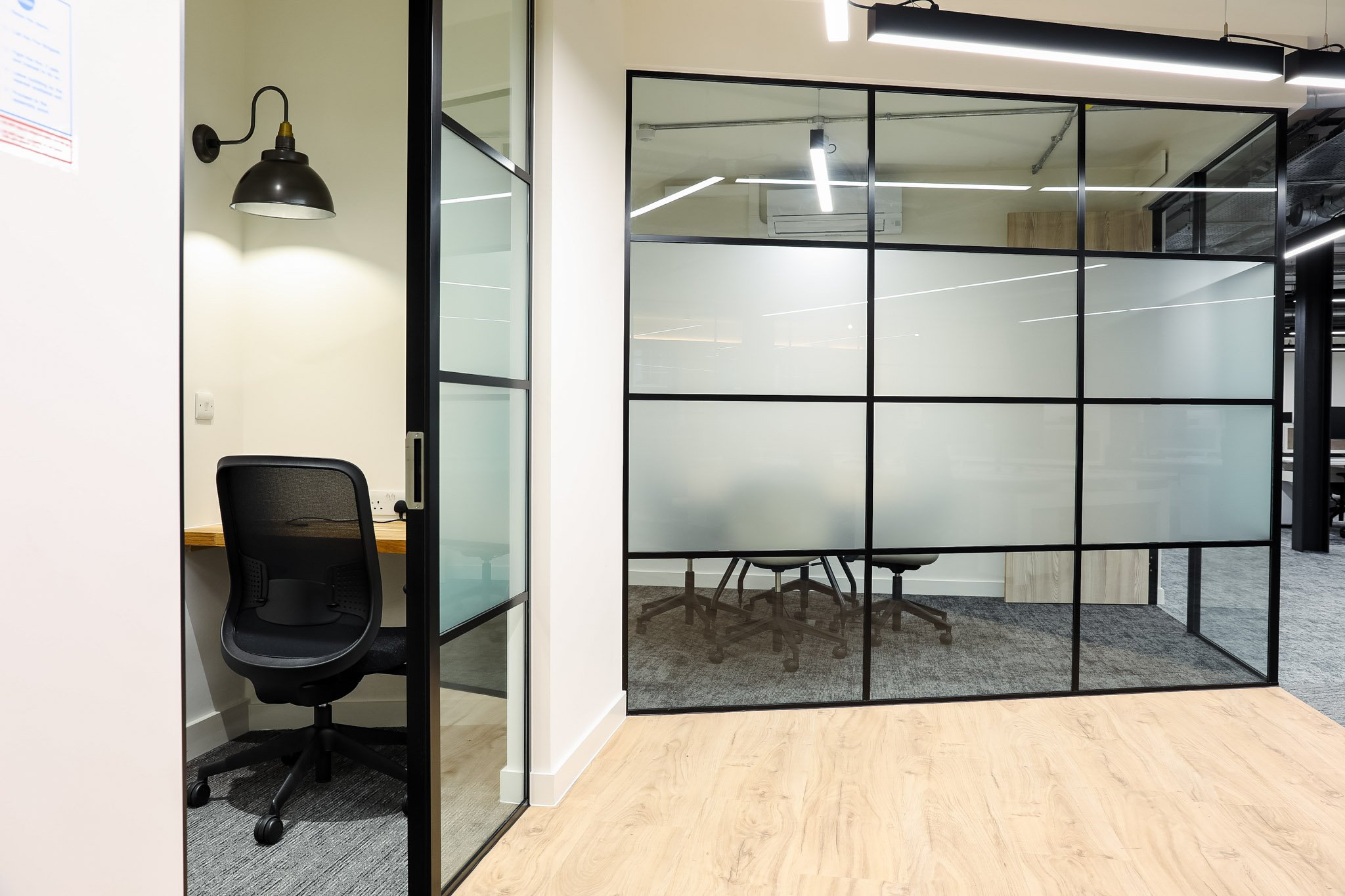 A modern office space with glass walls, a black office chair, and a small meeting area with a round table and office chairs. The space is well-lit with ceiling lights and a wall-mounted lamp.