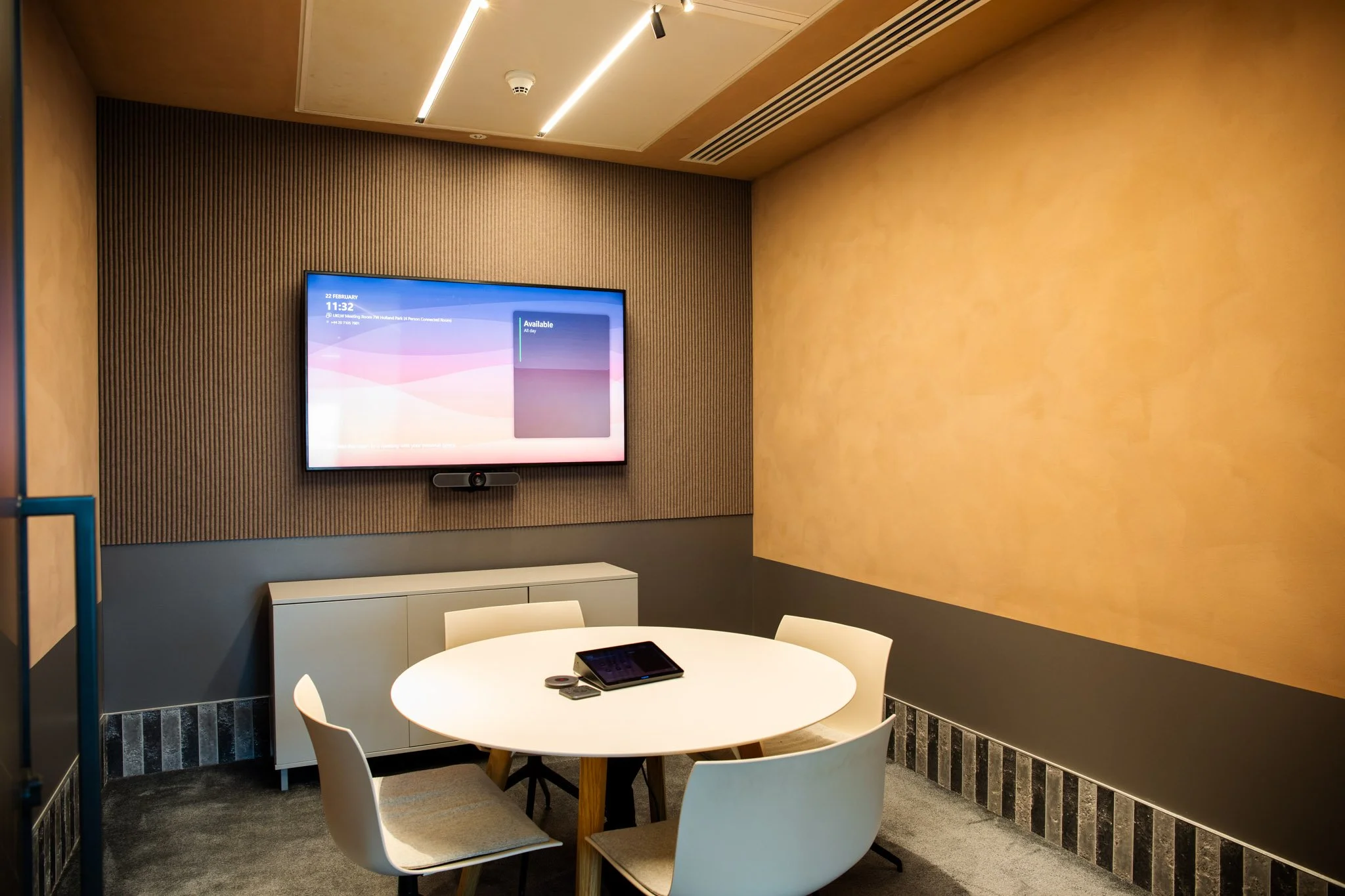 A modern conference room with a round white table and four white chairs. A digital screen on the wall displays the date, time, and availability information. A tablet and conference call device are on the table. The walls are decorated with textured a