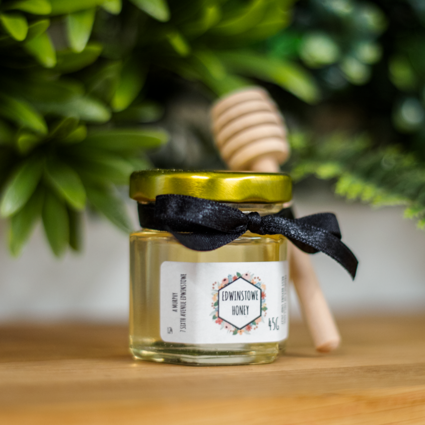 A jar of Edwistowe honey with a metal lid, decorated with a black ribbon, placed on a wooden surface with greenery in the background, and a wooden honey dipper leaning against the plant.