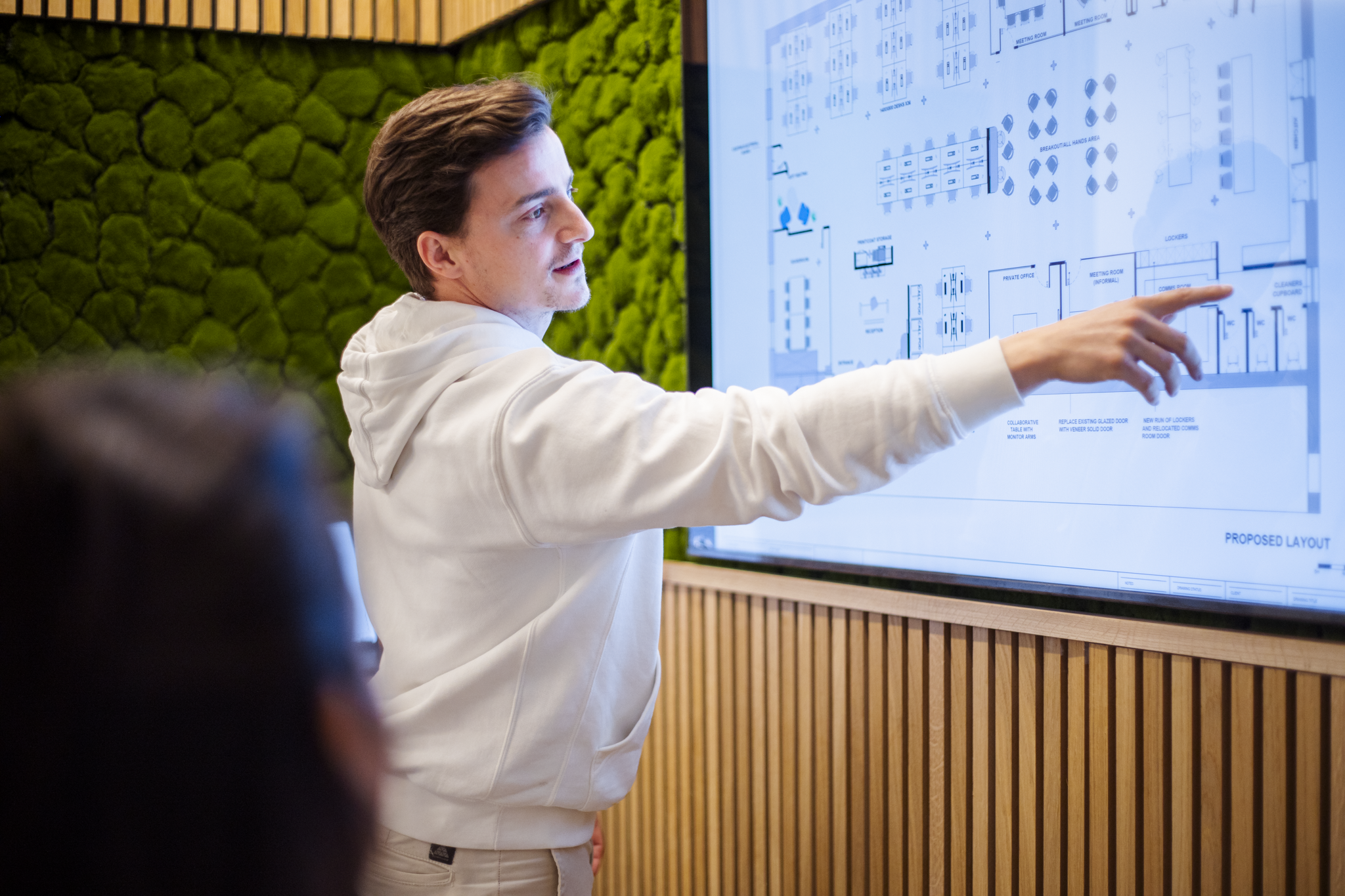 A man with brown hair in a white hoodie points at a large digital display with architectural diagrams, in a room with a green moss wall and wooden paneling.
