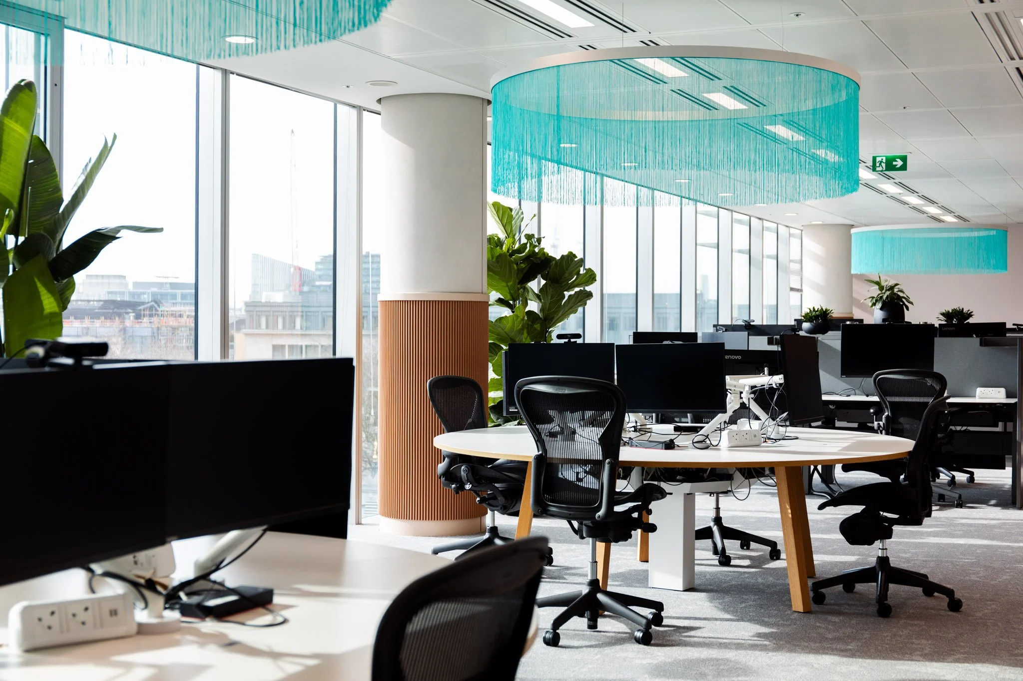 Modern office space with large windows, multiple computers and chairs, decorative plants, and large blue circular light fixtures hanging from the ceiling.