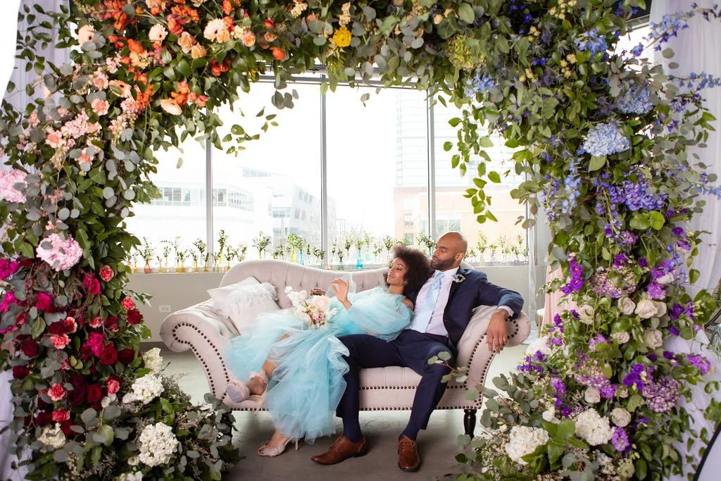 a couple sitting under a large floral arch on a pink couch