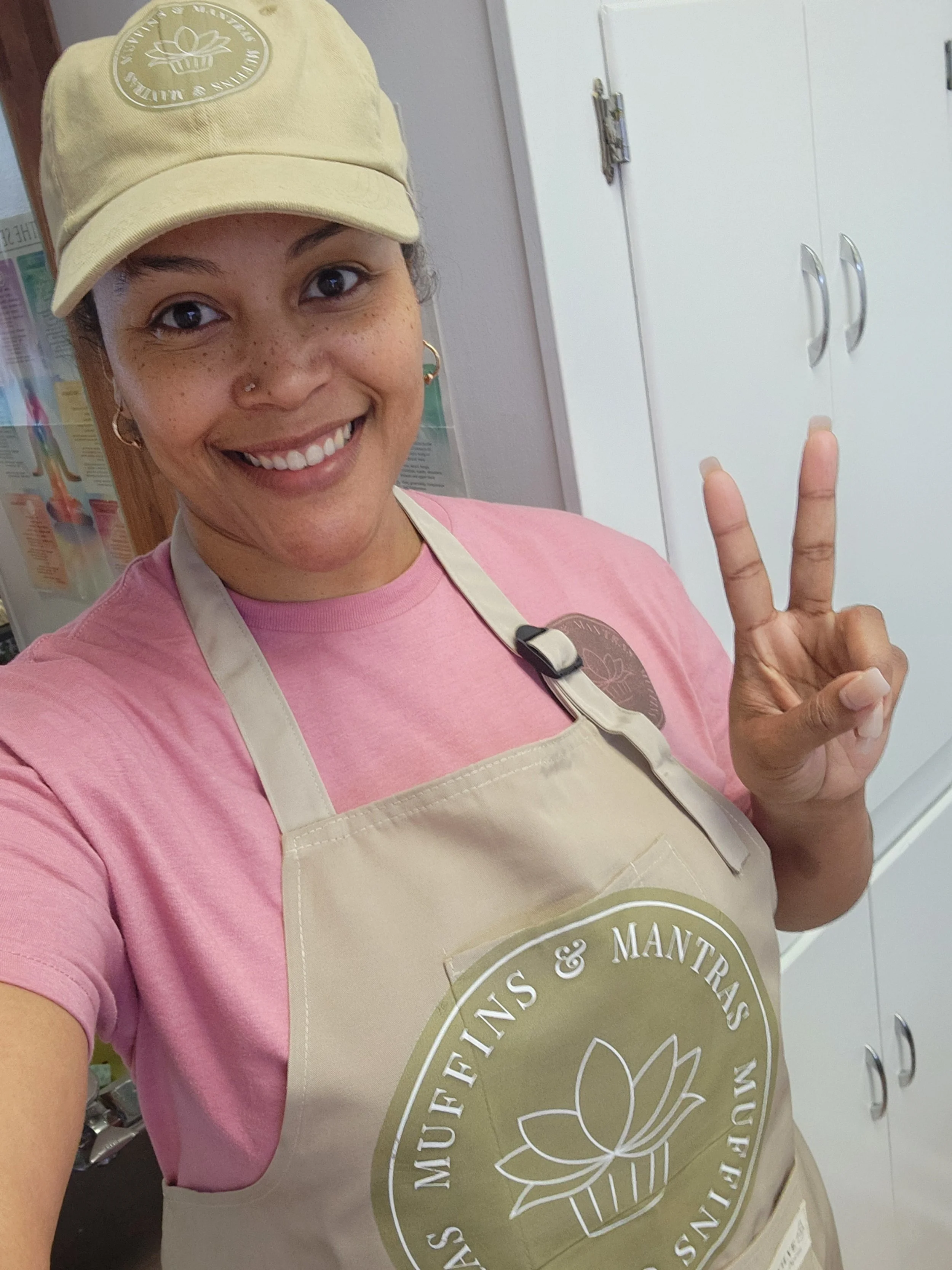 A woman taking a selfie, smiling, making a peace sign with her fingers in an indoor setting. She is wearing a beige apron with a logo, a pink t-shirt, and a beige cap. The background shows white cabinets and a poster with anatomical diagrams.