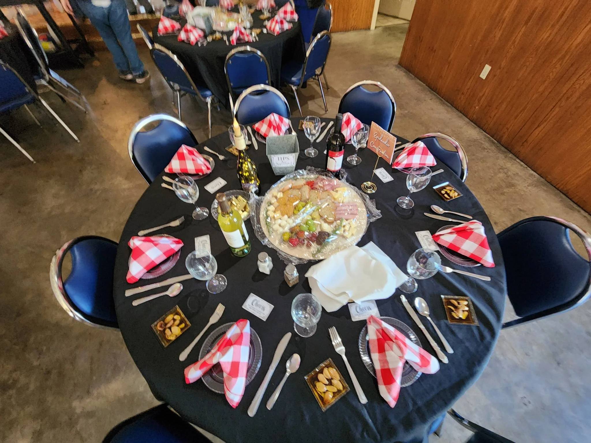 A round table set for a celebration, with red and white checkered napkins, wine glasses, and a large platter of cheese, crackers, and fruit in the center. Decorations include small signs, bottles of wine, and party favors.