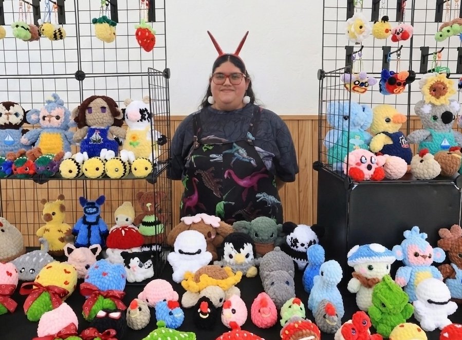 A woman with glasses and bunny ears headband displays crochet plush toys at a craft fair.