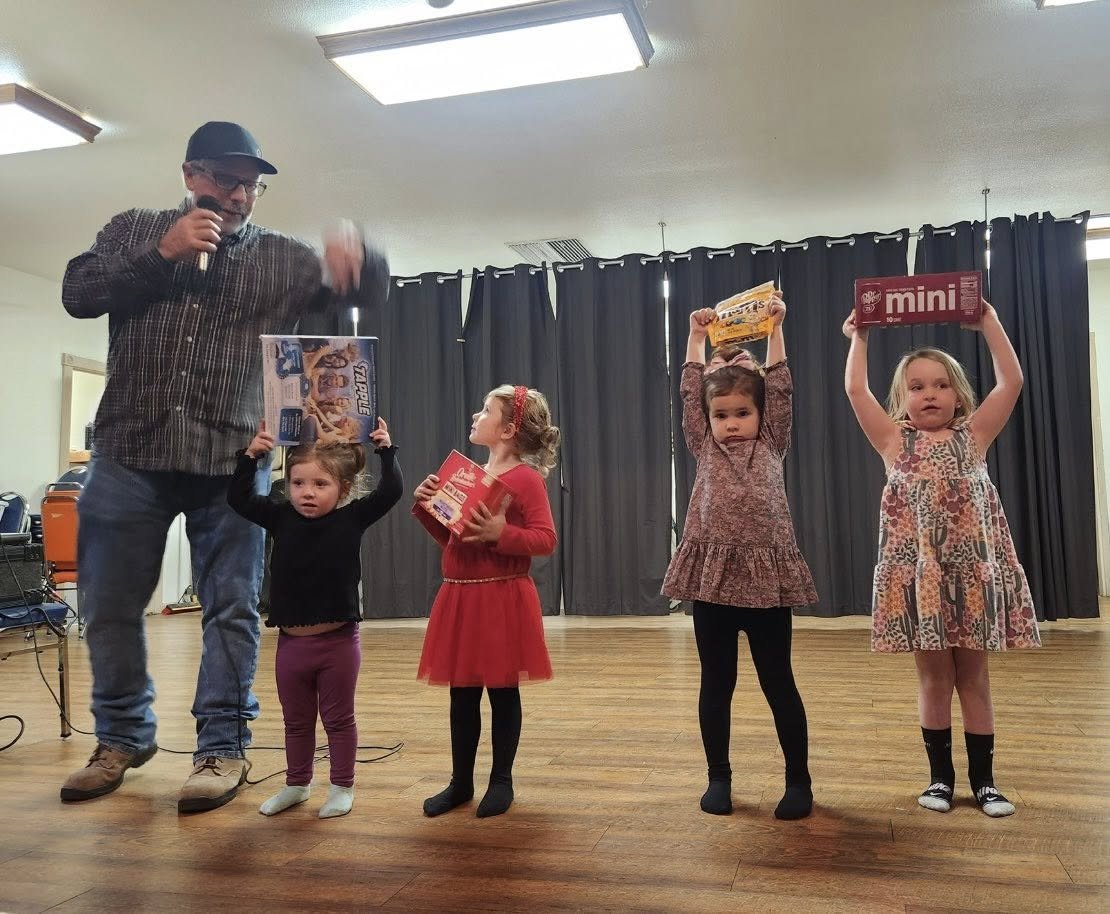 A man is standing on a stage with four young girls, all holding boxes above their heads. The man is speaking into a microphone. The girls are standing in a row with a dark curtain behind them and a wooden floor underneath. The girls appear to be rece