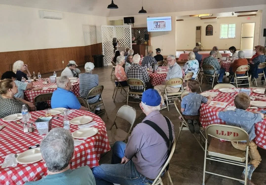 People seated at round tables with red checkered tablecloths in a banquet hall, attending a presentation or event, with a speaker and a TV screen at the front.