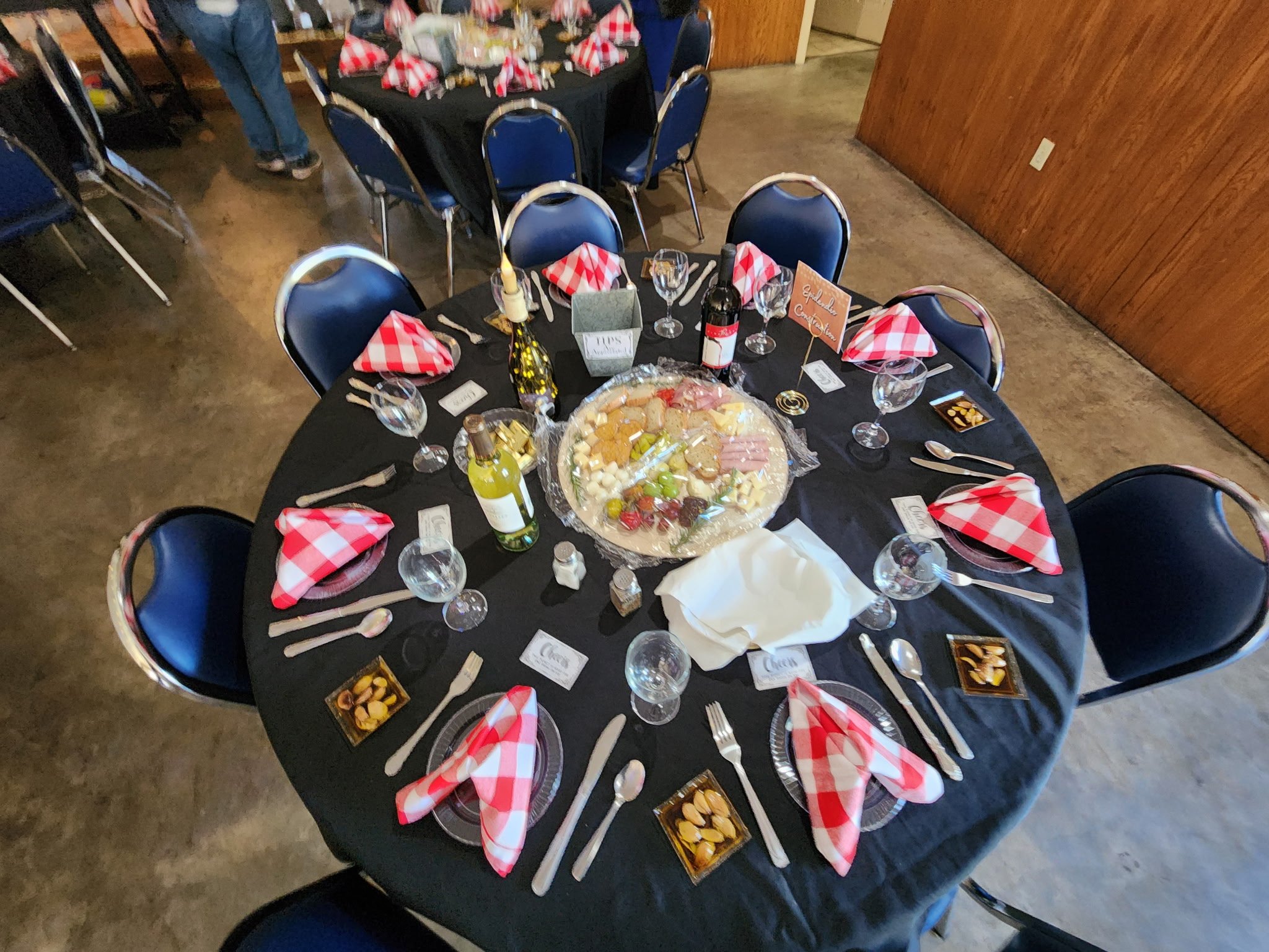 A round banquet table set for a celebration with black tablecloth, red checkered napkins, wine glasses, and a cheese platter in the center, in a room with wooden walls.