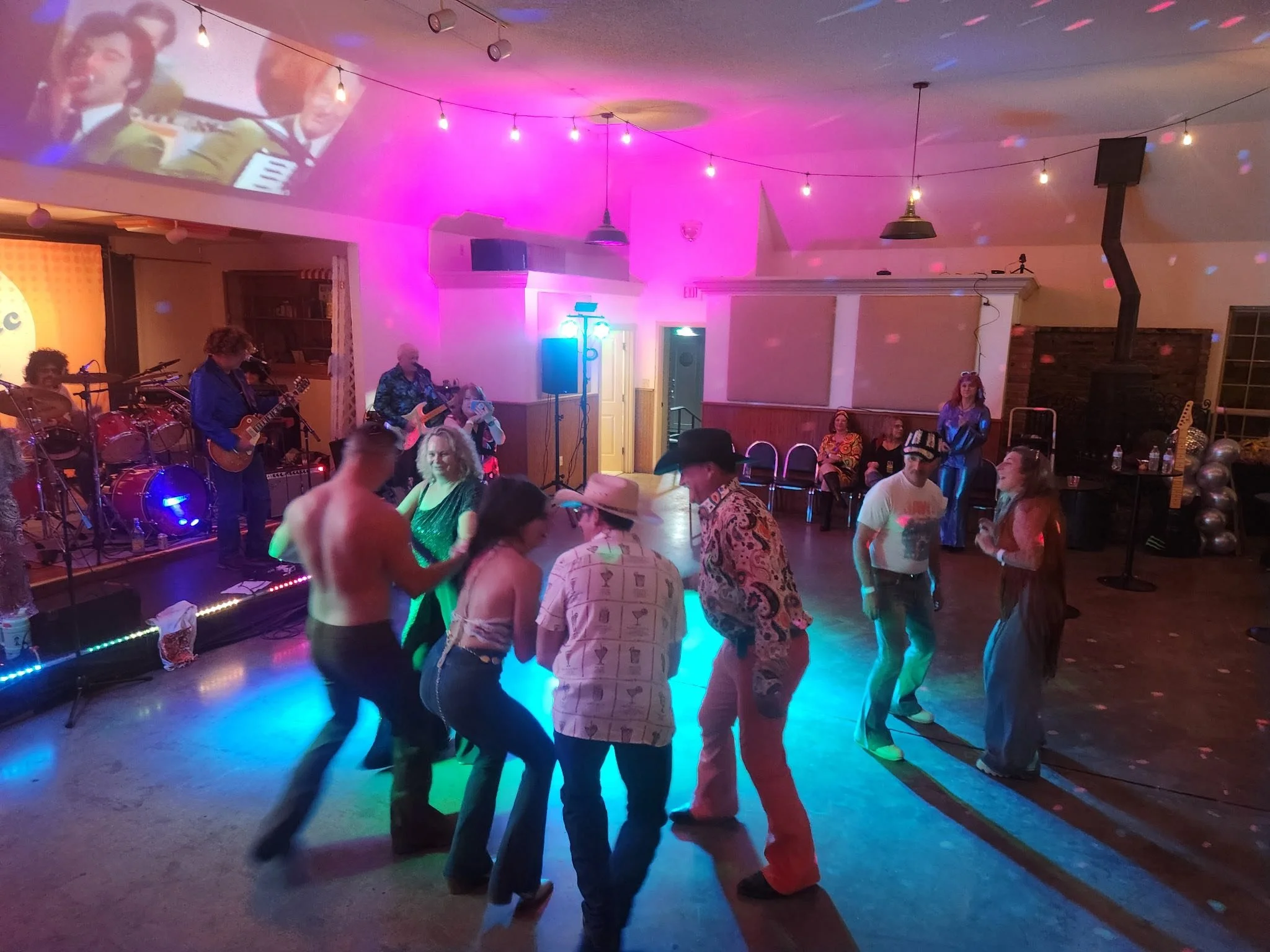 People dancing on a dance floor with a live band playing on stage, colored lights, and decorations in a lively indoor setting.