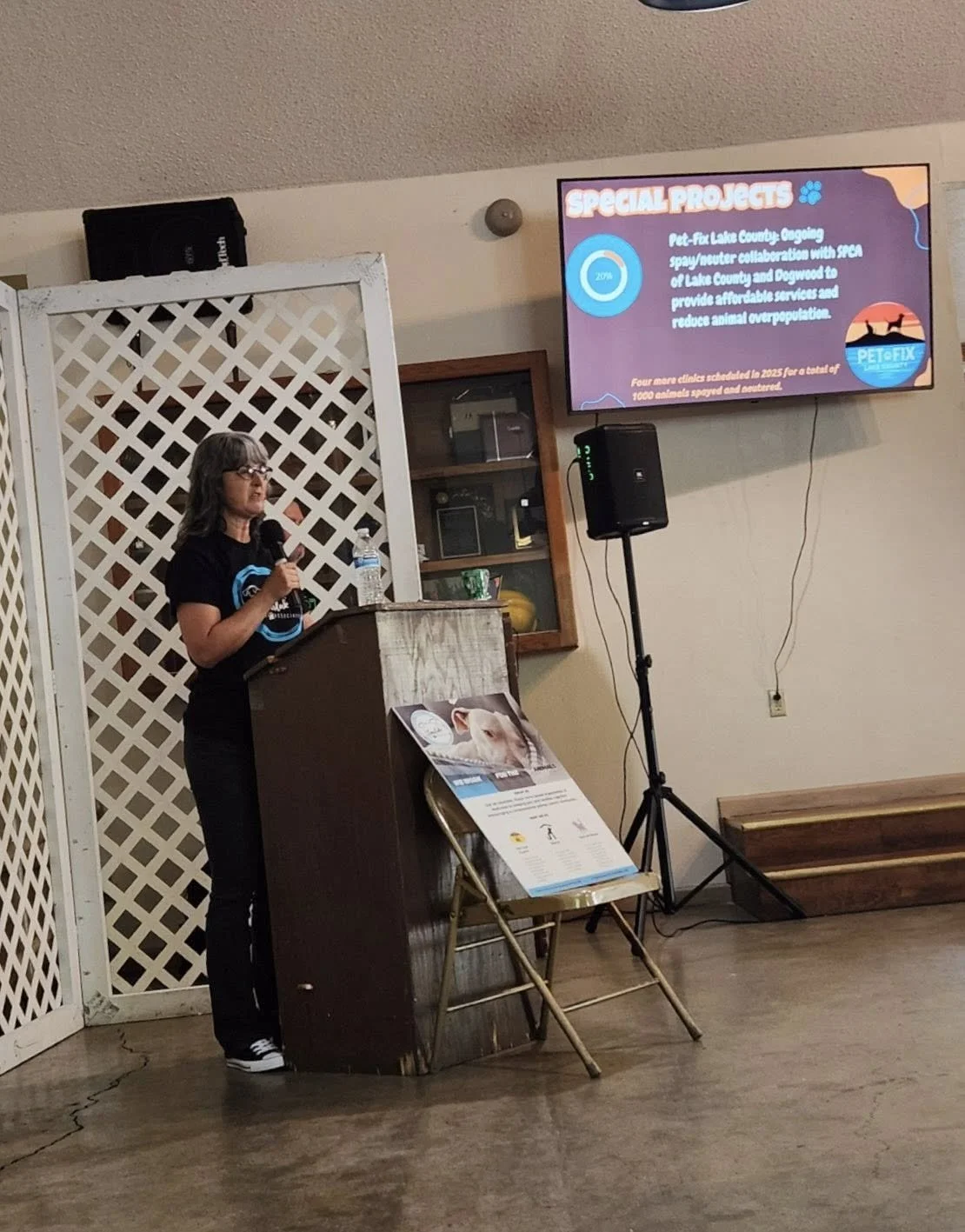 A woman giving a presentation at a podium with a microphone and a water bottle. Behind her is a lattice room divider, and a large display screen showing a slide about pet care projects for an animal organization.