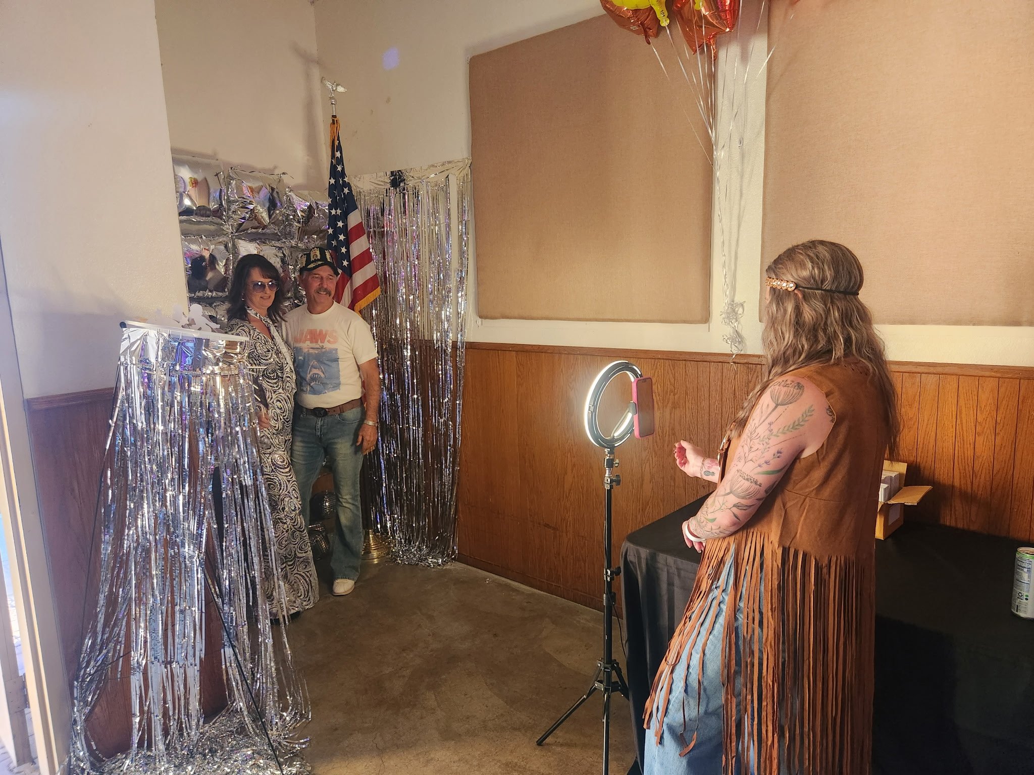 A woman with tattoos and long hair takes a photo of a couple standing in a decorated room. The couple is smiling, with the woman wearing sunglasses and a patterned dress, and the man in a white T-shirt and jeans. The background features silver tinsel