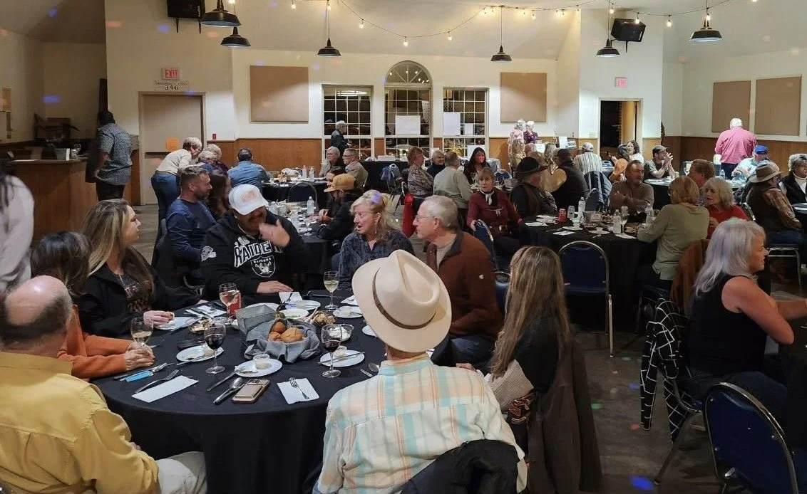 People gathered in a large dining hall with round tables, eating and socializing during an event. The room has string lights and a high ceiling with pendant lights.