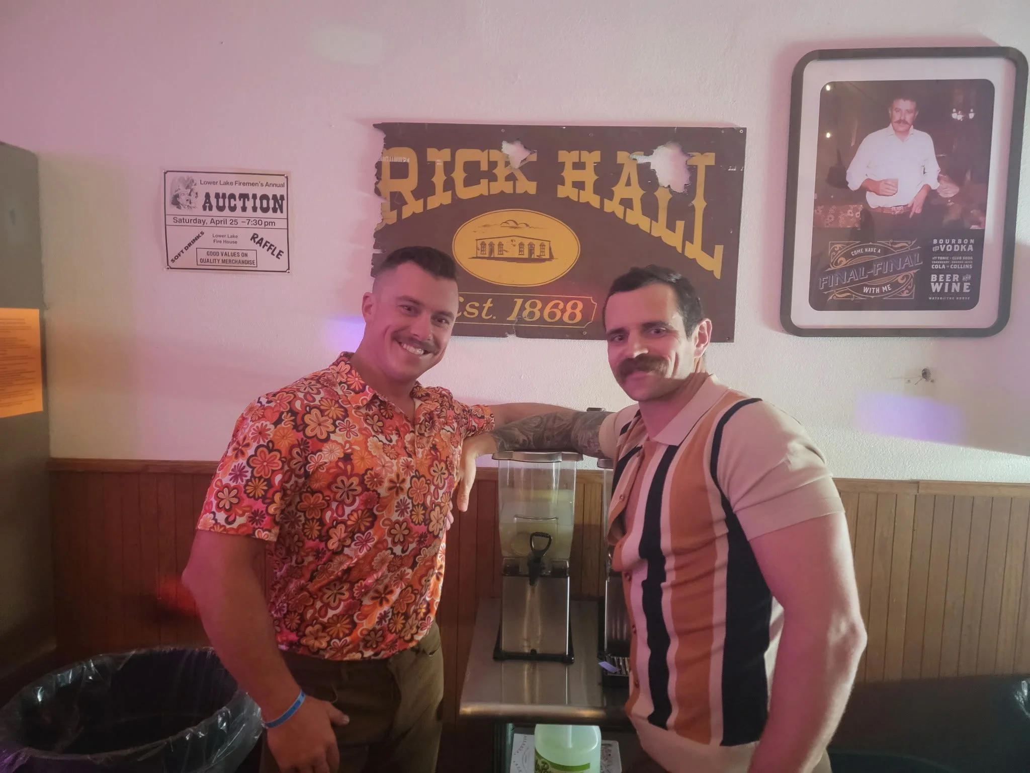 Two men stand indoors, smiling at the camera, with their arms resting on a water dispenser. The man on the left wears a colorful floral shirt, and the man on the right wears a striped beige and black shirt. Behind them are wall decorations including 