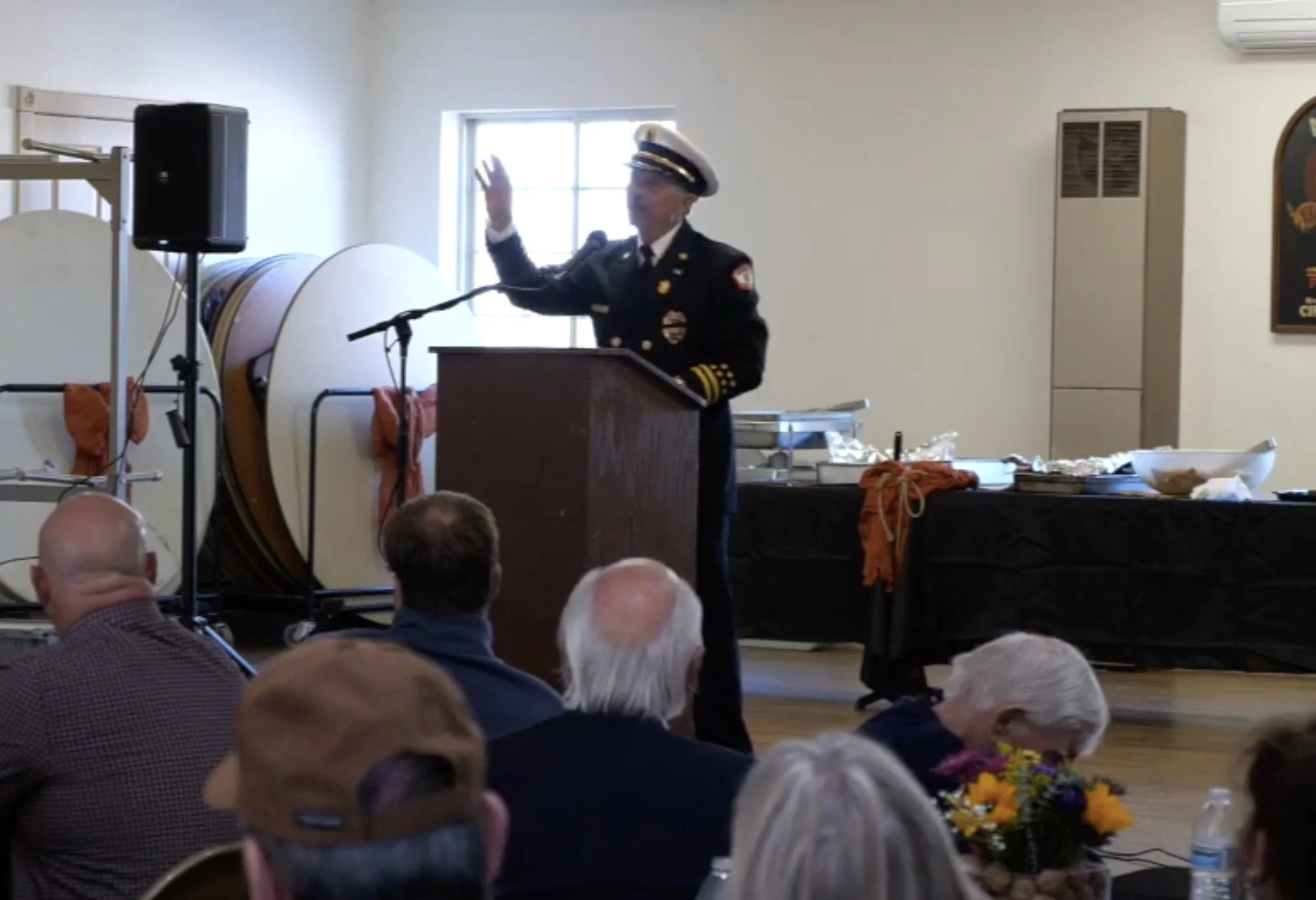 A firefighter in formal dress uniform giving a speech at a podium in front of an audience, with a table of food and a large drum in the background.