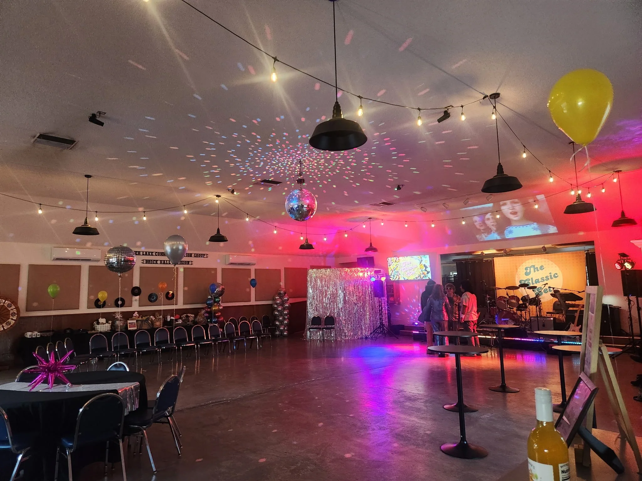 The image depicts a decorated event hall likely set up for a dance or celebration. The ceiling is adorned with string lights, hanging disco balls, and a yellow balloon. The dance floor is empty, with a few groups of people standing and talking near t