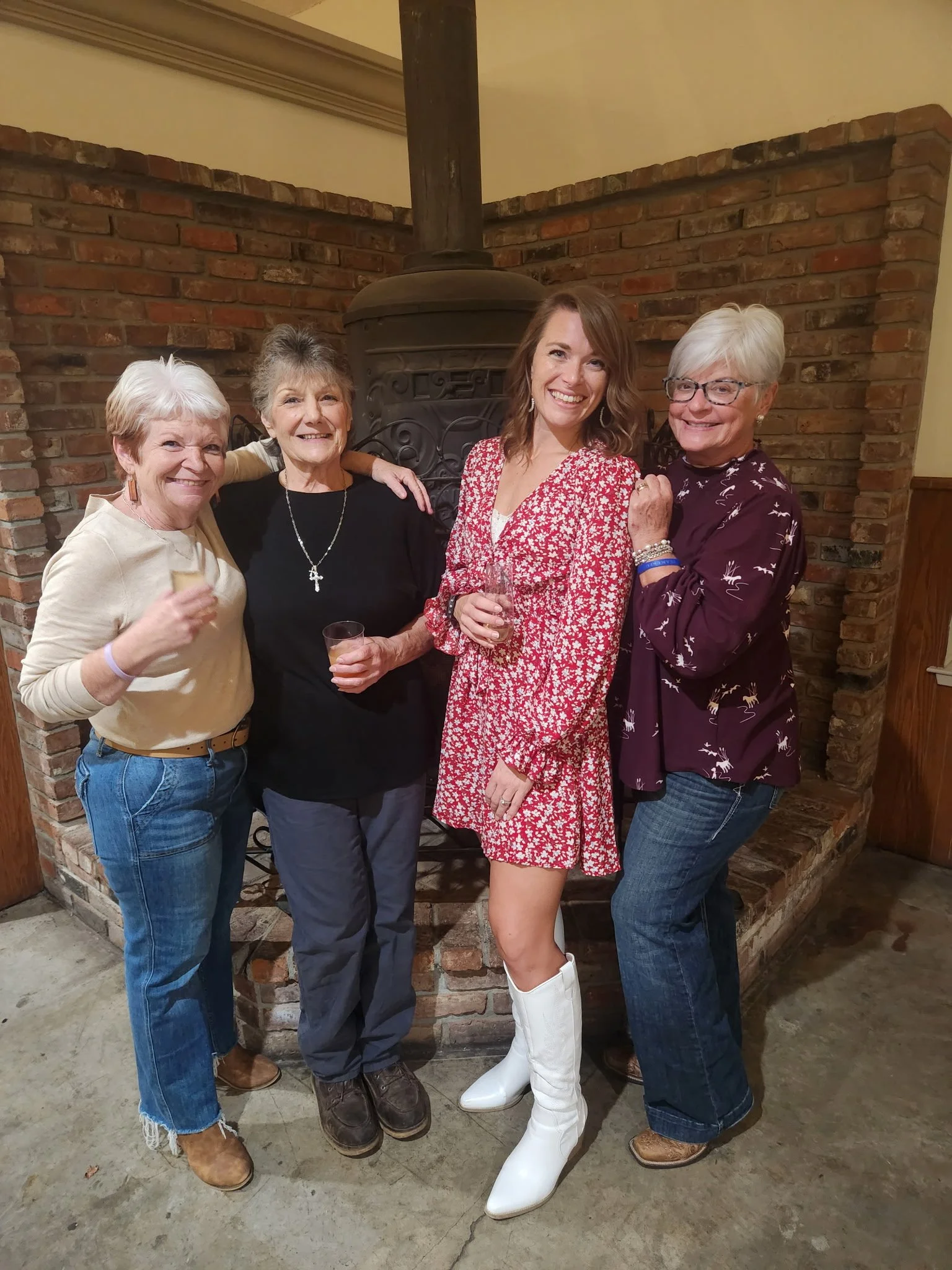 Four women standing close together in front of a brick fireplace, smiling, with drinks in hand.