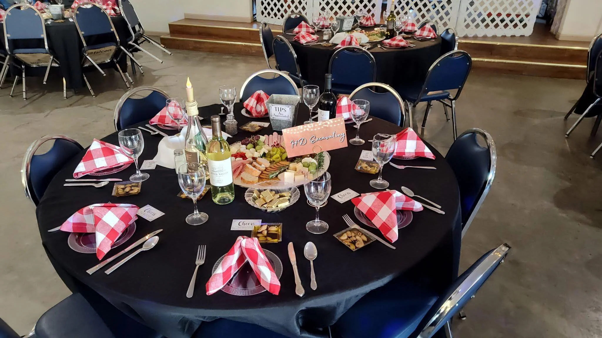 A round banquet table set for a formal event with a black tablecloth, red and white checkered napkins, wine glasses, cutlery, and a centerpiece of cheese, crackers, grapes, and a decorative candlestick. There are place cards and a sign indicating it 