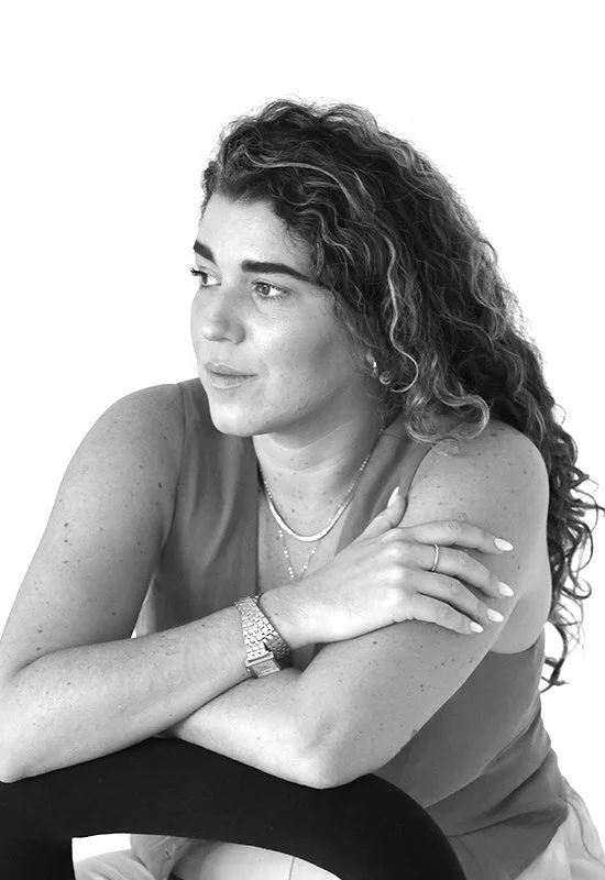 A woman with curly hair and light skin, sitting with her arms crossed, looking to the side, in a sleeveless top, wearing jewelry and a watch, against a plain white background.