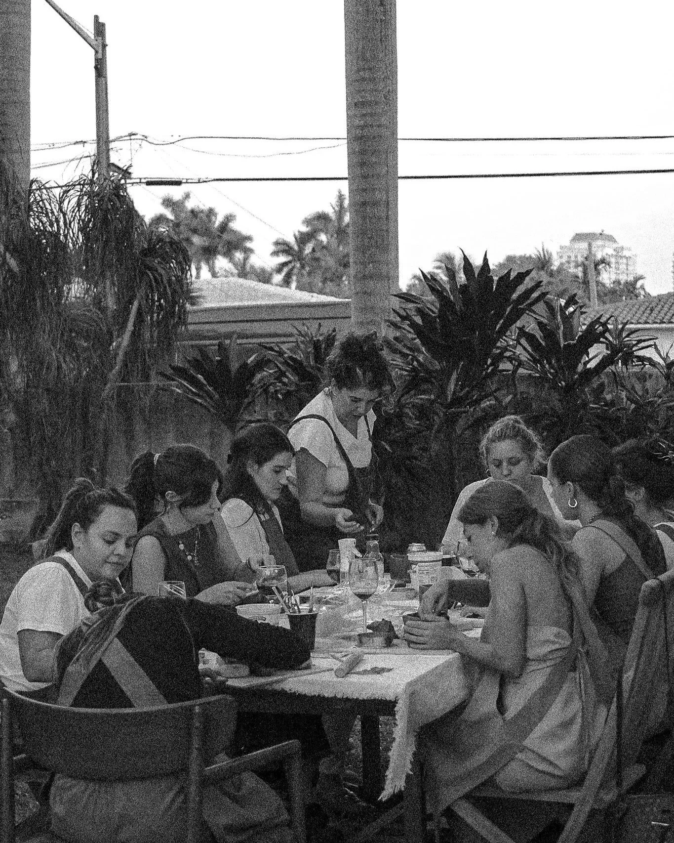 A group of women sitting around a table outdoors, engaged in a crafting activity, with lush plants and electrical wires in the background.