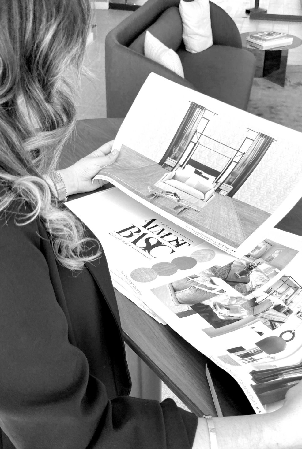 A woman sitting at a table reviewing a furniture catalog, with a living room area in the background.