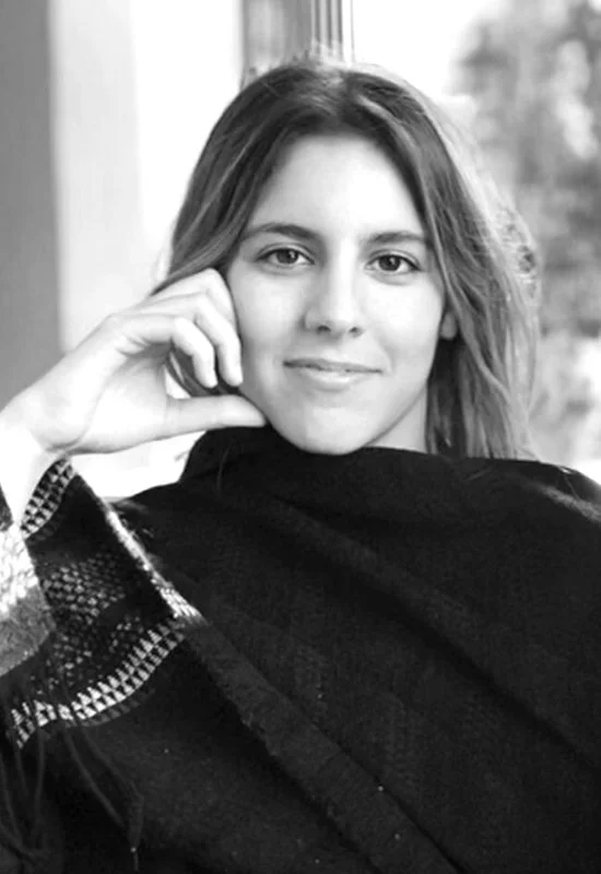 Black and white photograph of a young woman with long hair, smiling slightly, sitting indoors near a window with blurred outdoor scenery, wearing a dark shawl over a checkered shirt, resting her head on her hand.