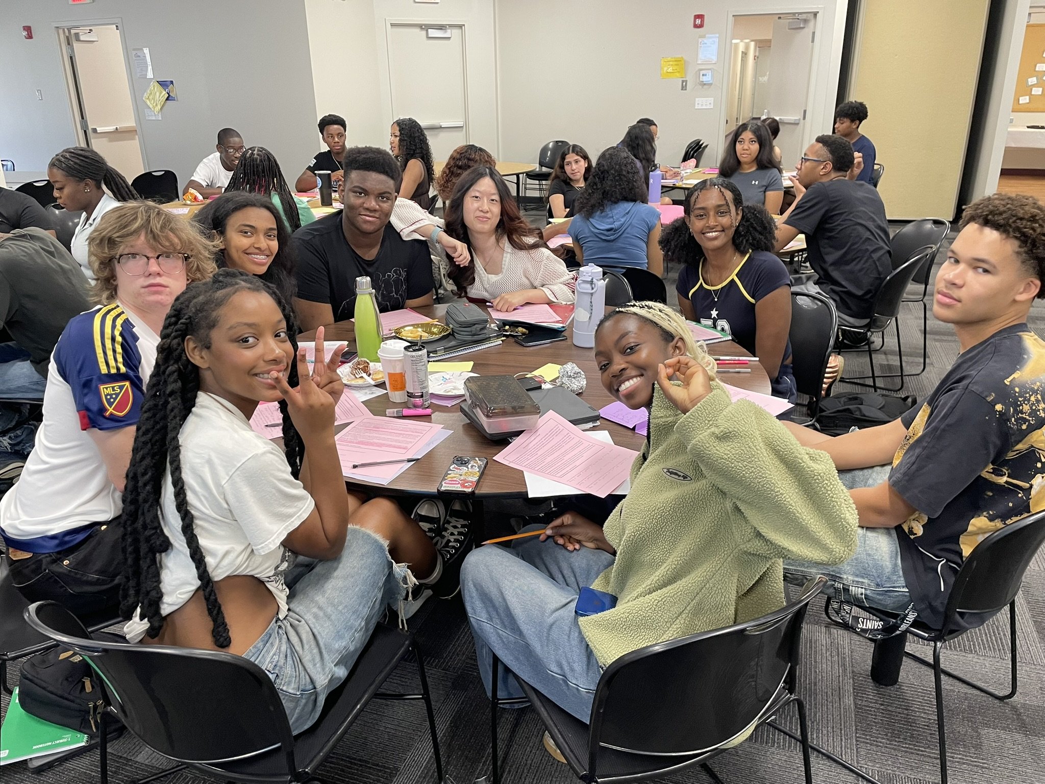 A group of diverse young people sitting around a table in a classroom or conference room, engaging in a discussion or activity, with papers, notebooks, and drinks on the table.