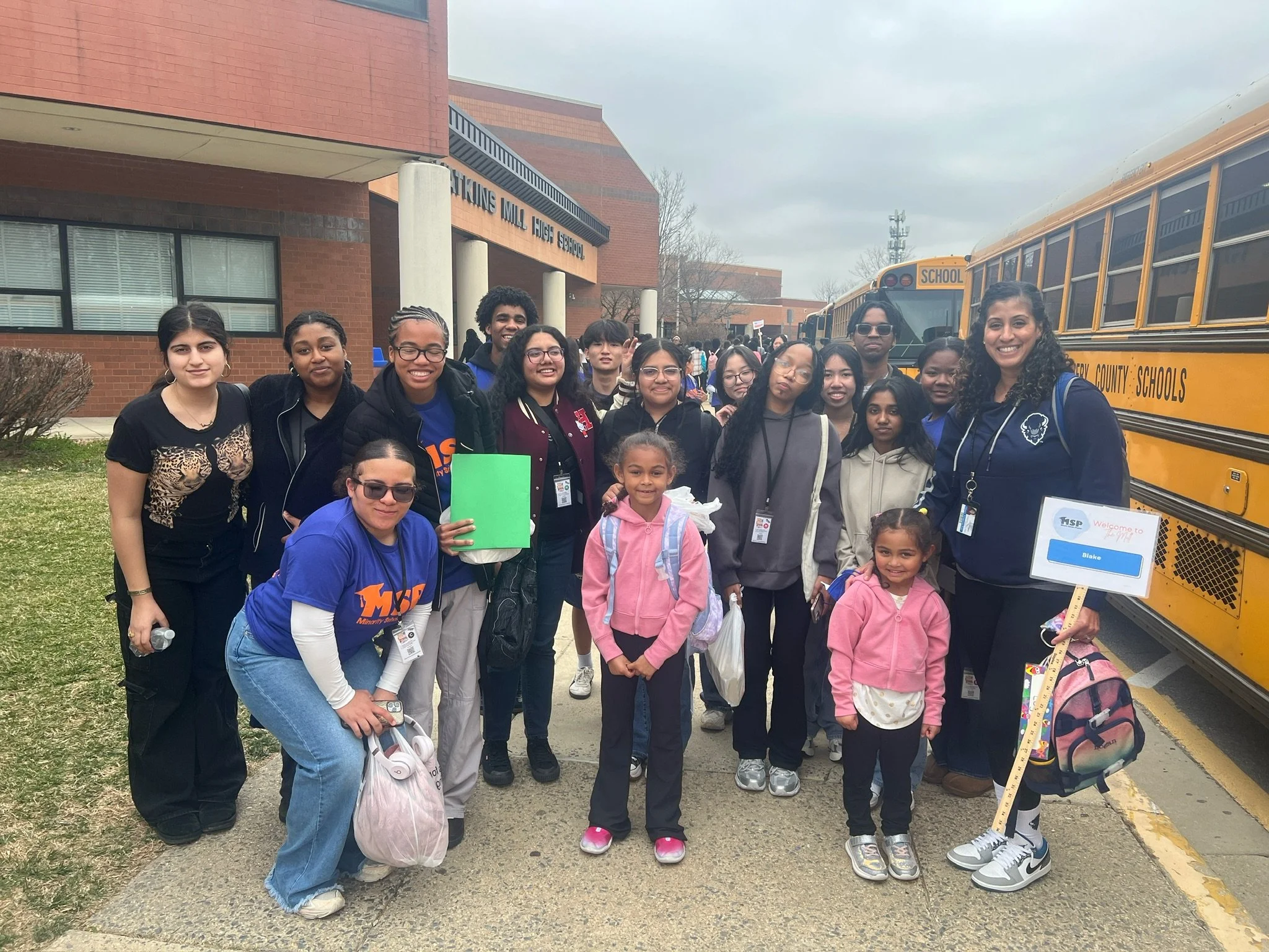 Group of diverse students and teachers standing outside school with yellow buses in the background.