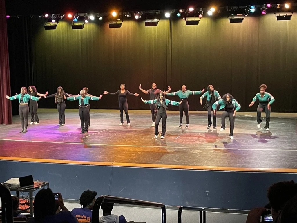 A group of performers on stage, wearing matching teal and black outfits, dancing with arms extended in various directions under stage lights, with an audience watching.