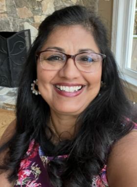 A smiling woman wearing glasses and earrings, with long dark hair, sitting indoors near a window with a stone fireplace in the background.