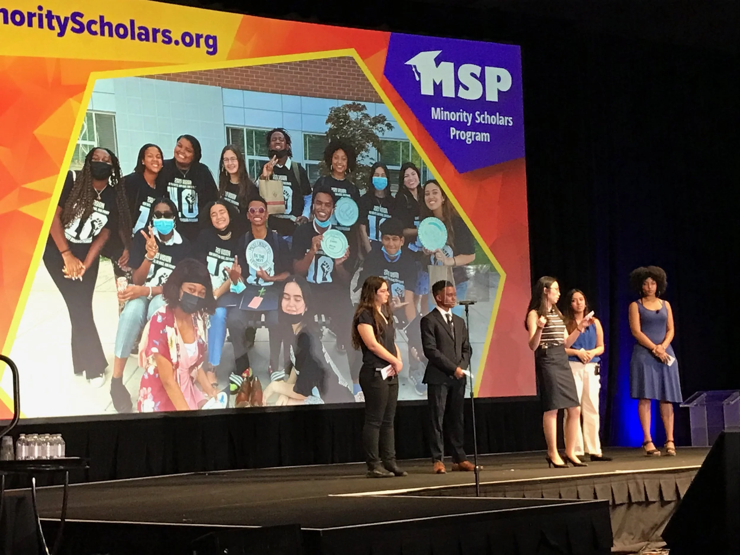 Group of young people on stage at an awards ceremony, with a large screen behind them displaying a photo of a diverse group of students holding medals and trophies, for the Minority Scholars Program.