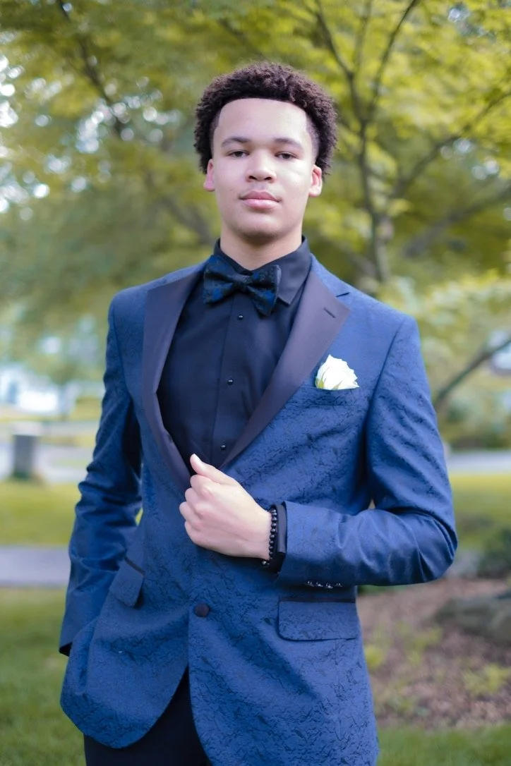 A young man in a blue textured tuxedo with black lapels and a matching bow tie, standing outdoors with green trees in the background. He has curly hair and is holding his tuxedo jacket with one hand, wearing a black bracelet on his wrist, and has a white pocket square in his jacket pocket.