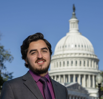 A man with dark hair and a beard smiling in front of the U.S. Capitol building in Washington, D.C.