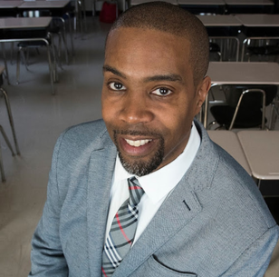 Close-up of a man in a gray suit with a plaid tie, smiling in a classroom or conference room setting.