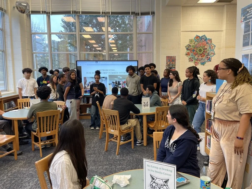 A group of students gathered around a presentation in a library media center, with a person speaking in front of a large screen displaying a summer internship program.